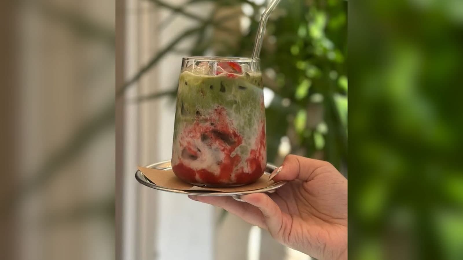 Elde tutulan, cam bardakta çilekli matcha latte; arkada yeşil bitkiler
